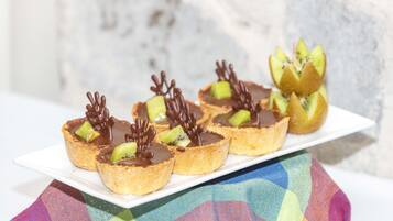 A white rectangular plate with six chocolate-topped pastries, each garnished with kiwi slices and chocolate decorations, placed on a colorful checkered cloth.