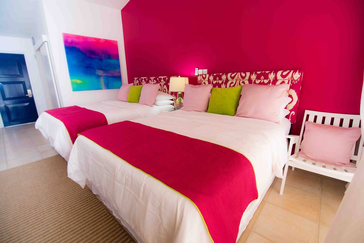 Une chambre d’hôtel avec deux lits, un mur rouge, un tableau et une chaise blanche.