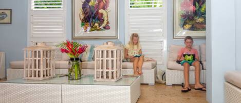 A room with a white wicker sofa, a glass coffee table with a vase of flowers, and framed botanical prints on the wall.