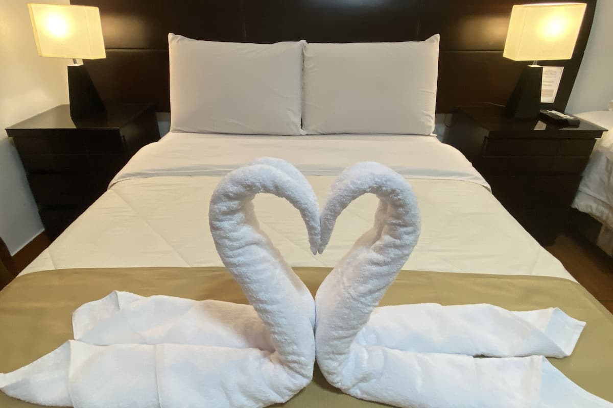 A hotel room with a neatly made bed, two bedside lamps, and a heart-shaped towel arrangement on the bed.
