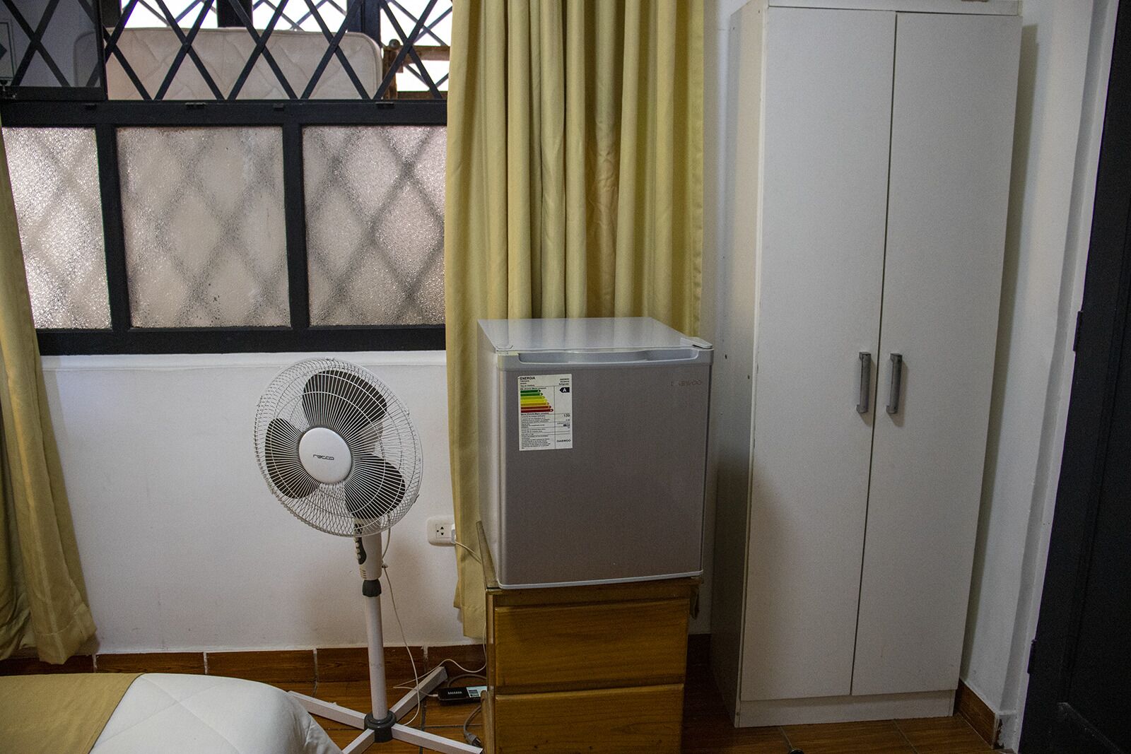 Ein Zimmer mit einem weißen Schrank, einem kleinen Kühlschrank, einem Standventilator und einem Fenster mit einem Metallgitter.