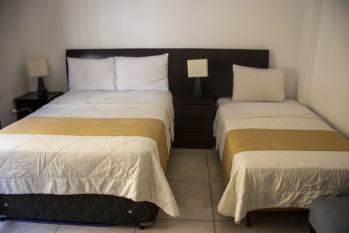 A hotel room with two beds, each with a duvet and pillows, a nightstand with a lamp, and a suitcase on the floor.