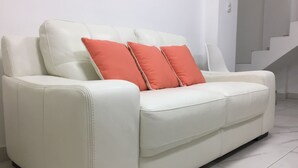A white leather sofa with two orange cushions in a modern, minimalistic room with white walls and a tiled floor. - Plaza Mayor Lima (Lima)
