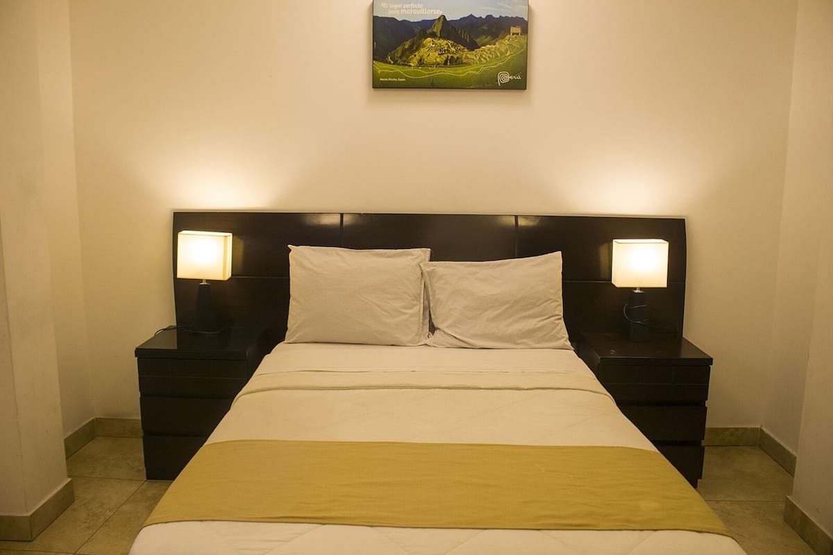 A neatly made bed with white linens and a yellow throw, flanked by two bedside tables with lamps, in a room with a framed landscape painting above the bed.