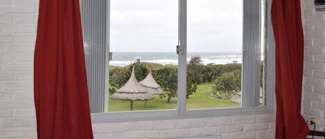 Una habitación con un amplio ventanal que da a un jardín con una estructura de techo de paja y el mar.