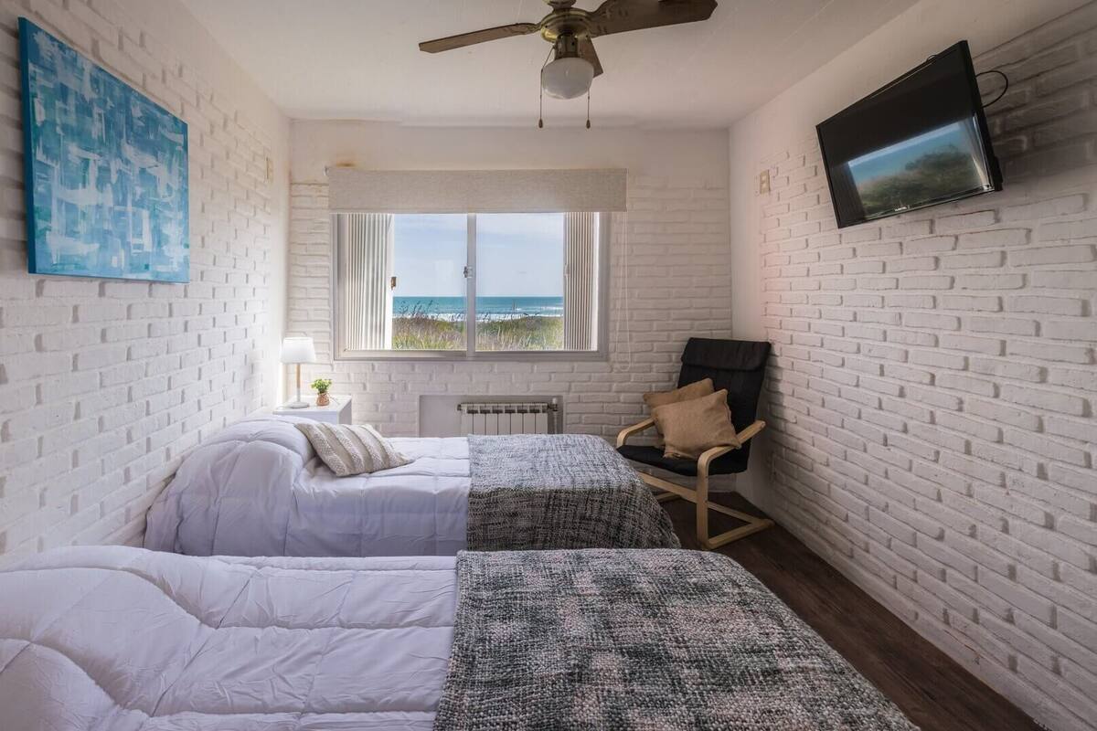 A room with two beds, a chair, a TV, and a window with a view of the sea.