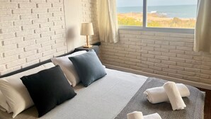 Habitación de hotel con pared de ladrillo, una cama con varias almohadas, una mesita de noche con lámpara y vista a la playa por la ventana.