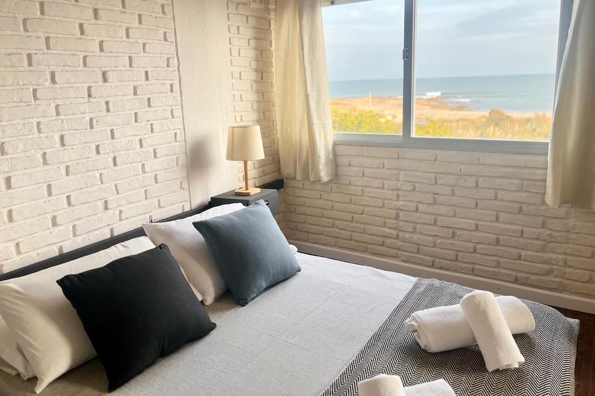 A hotel room with a brick wall, a bed with multiple pillows, a nightstand with a lamp, and a view of the beach through a window.