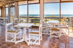 Un área de comedor al aire libre con mesas y sillas blancas, grandes ventanales con vistas al océano y una cerca de madera al fondo.