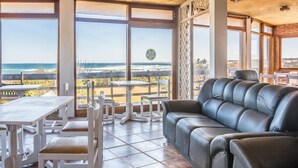 Amplio espacio interior con un gran ventanal que ofrece vista a la playa y al océano. Hay un sofá de cuero negro y muebles de madera blanca.