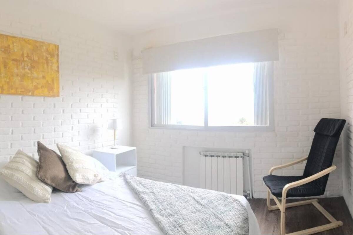 A bedroom with a white brick wall, a bed with pillows, a nightstand, a chair, a window, and a radiator.