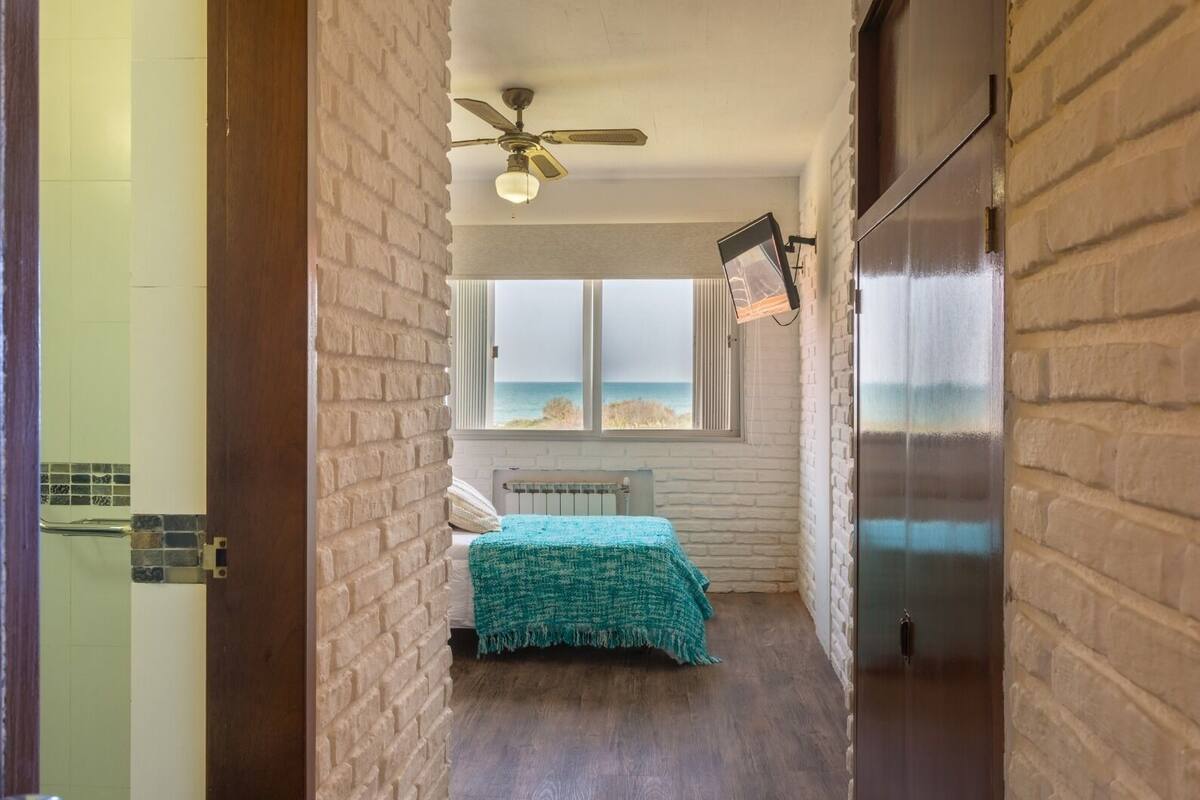 A narrow hallway leading to a bedroom with a bed, a ceiling fan, a window with a view of the beach, and a refrigerator.