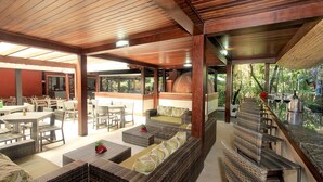Área de pátio externo com móveis de vime, forno de tijolos e guarda-corpo de vidro com vista para um jardim.