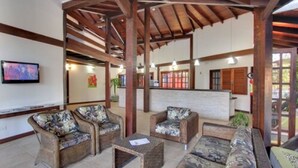 Um hall amplo com móveis de vime, vigas de madeira e uma televisão na parede.