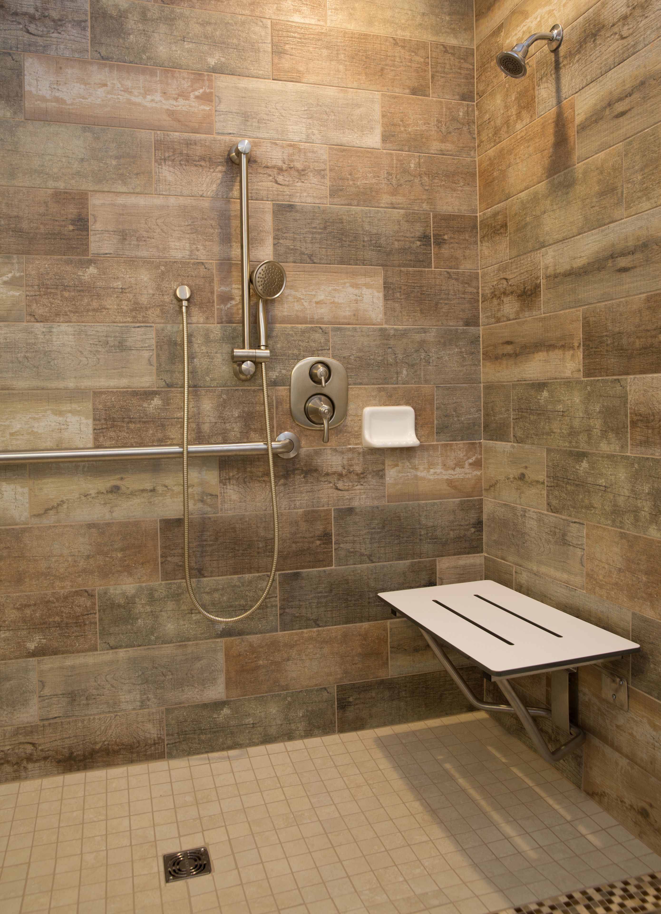 A shower area with a tiled wall, a handheld showerhead, a wall-mounted showerhead, a handheld shower hose, a soap dish, and a white bench.