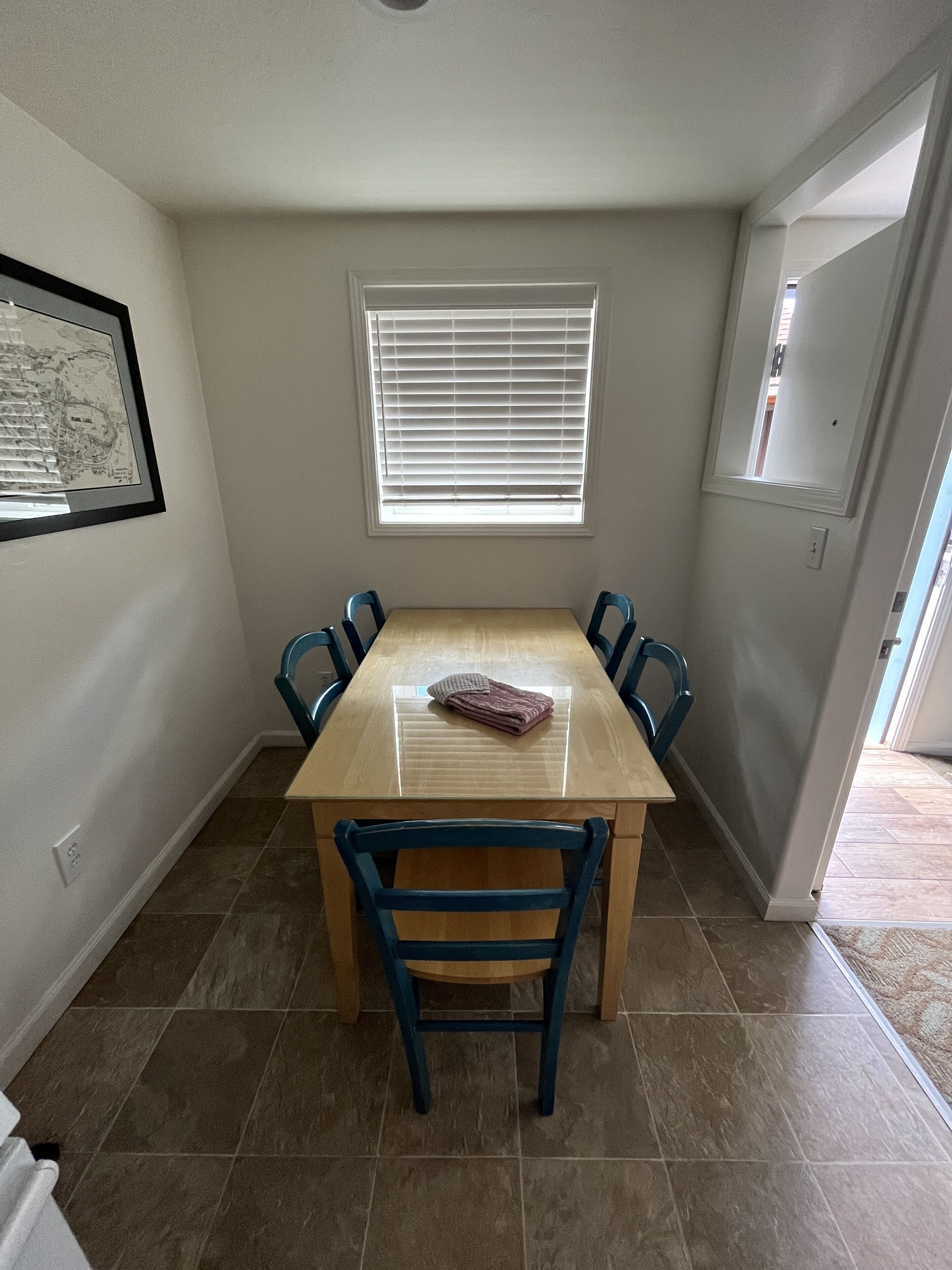 A dining area with a wooden table and blue chairs, a window with blinds, and a framed map on the wall.