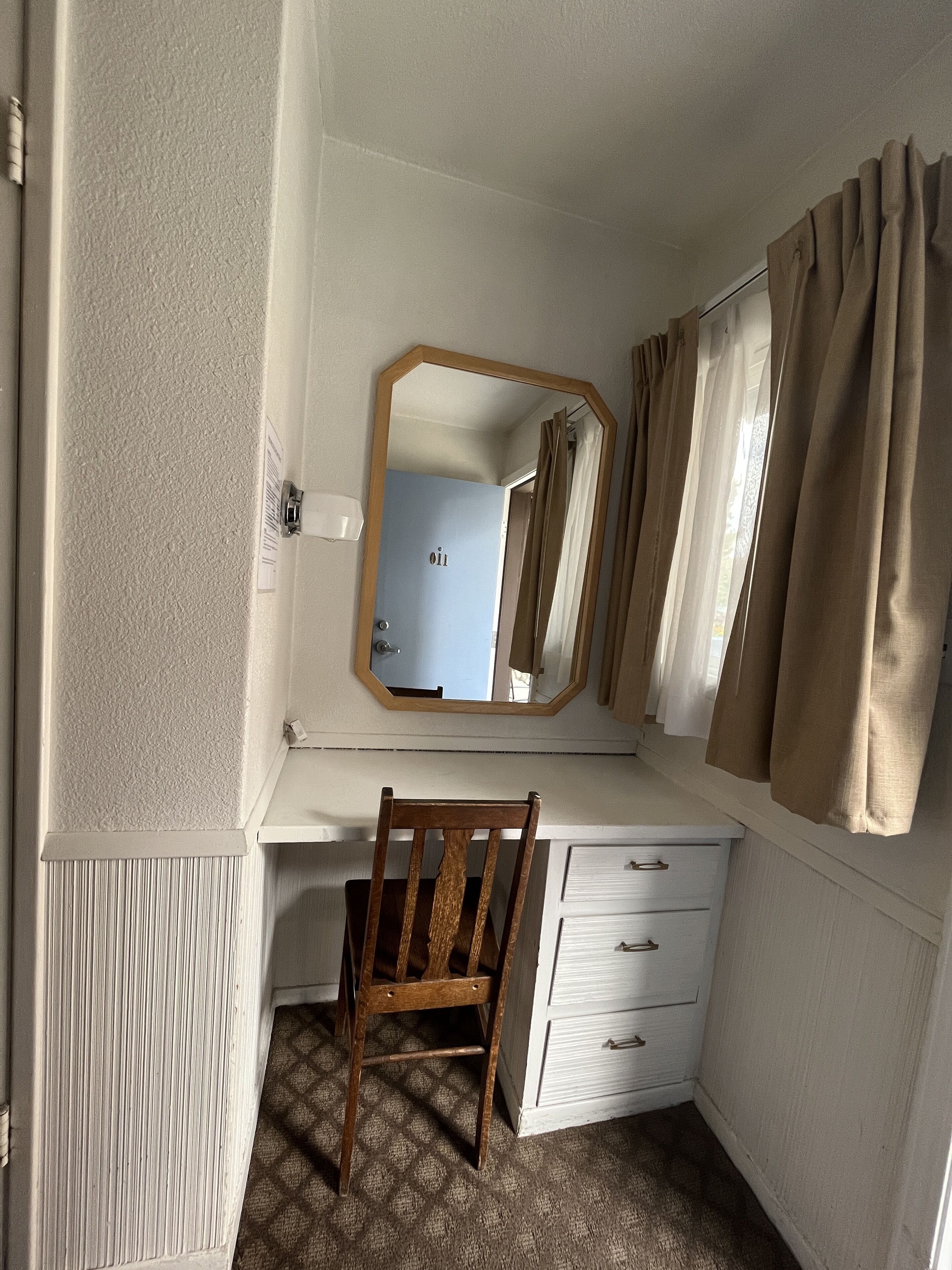 Une pièce de taille modeste, avec une chaise en bois, un bureau blanc, un miroir et une fenêtre avec des rideaux.