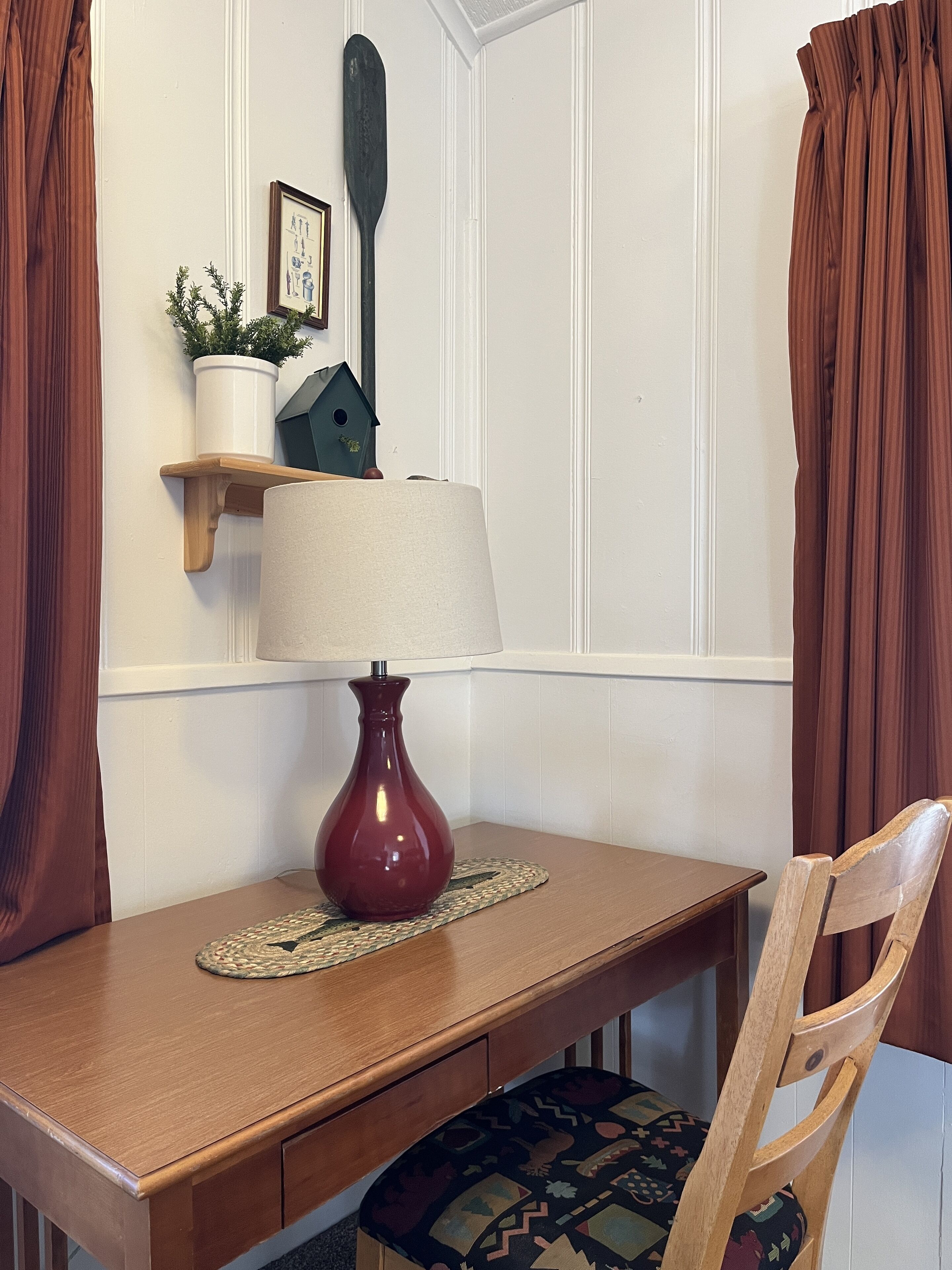 Una mesa de madera con una lámpara roja, un posavasos tejido y una silla con un cojín estampado.