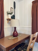 Una mesa de madera con una lámpara roja, un posavasos tejido y una silla con un cojín estampado.