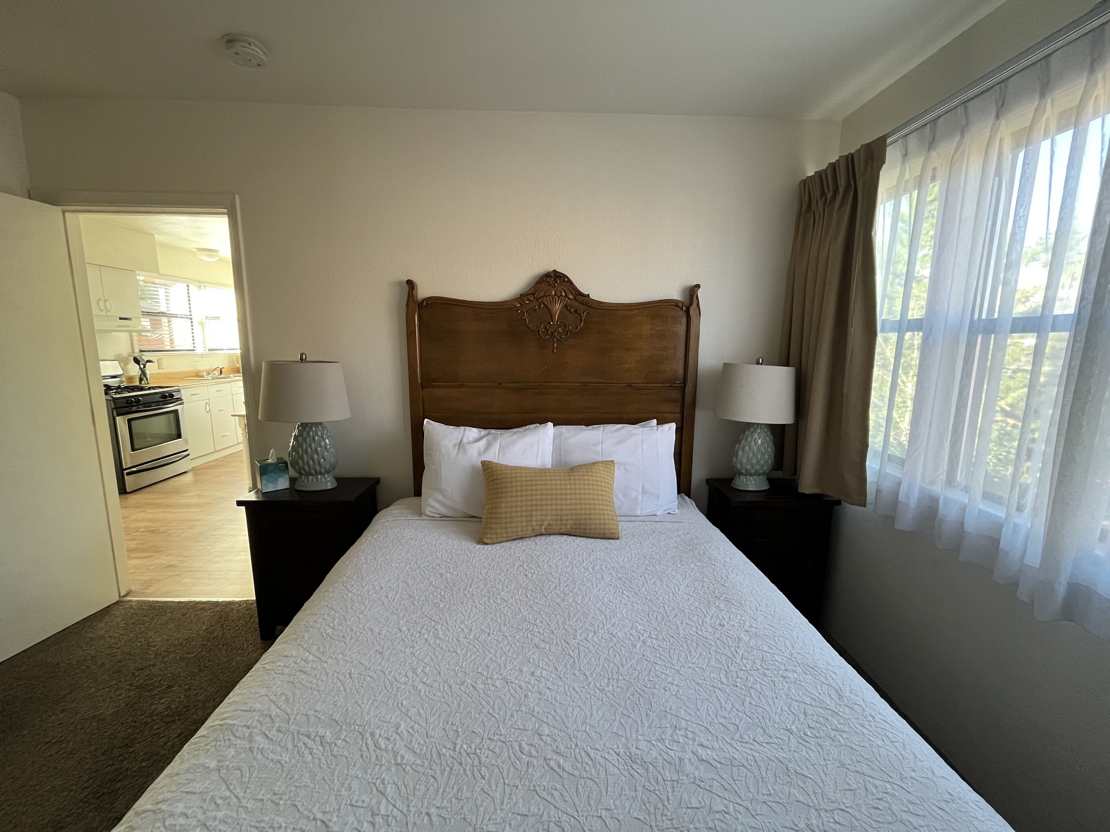 A hotel room with a large bed, wooden headboard, two bedside tables with lamps, and a view of the kitchen through an open door.