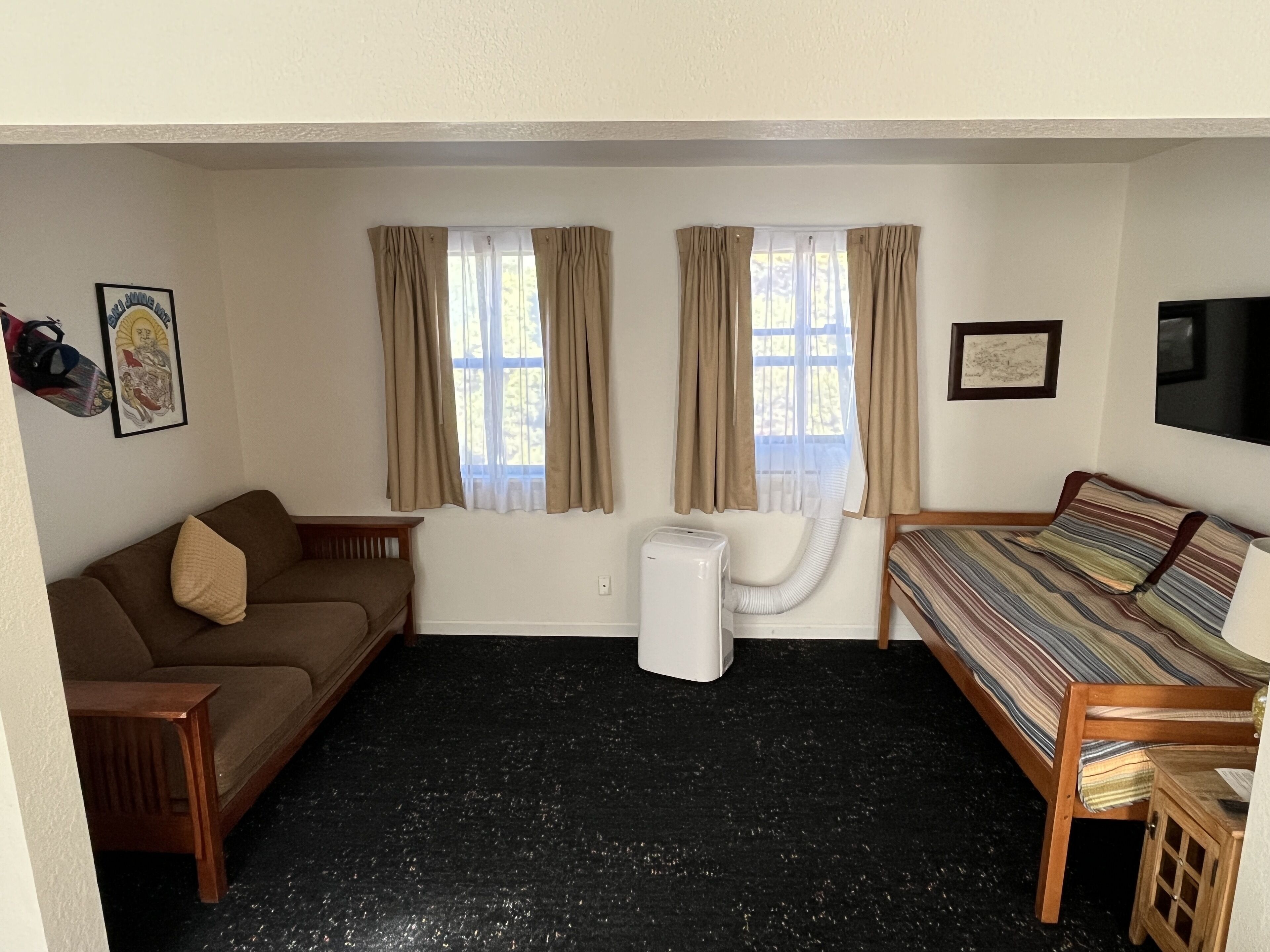 Una habitación con un sofá marrón, una cama a rayas, un calentador blanco y dos ventanas con cortinas color beige.
