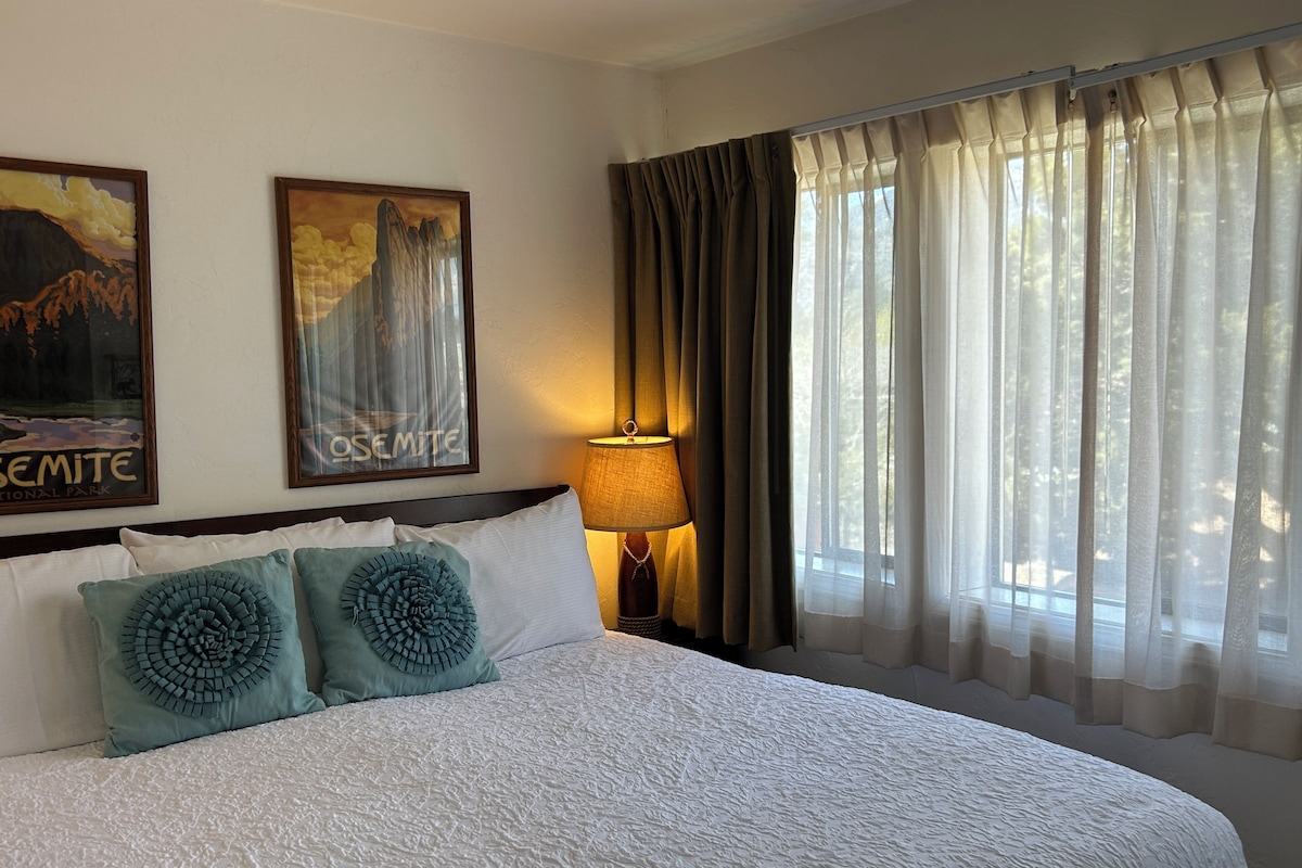 A hotel room with a large bed, two decorative pillows, a nightstand with a lamp, and curtains on the window.