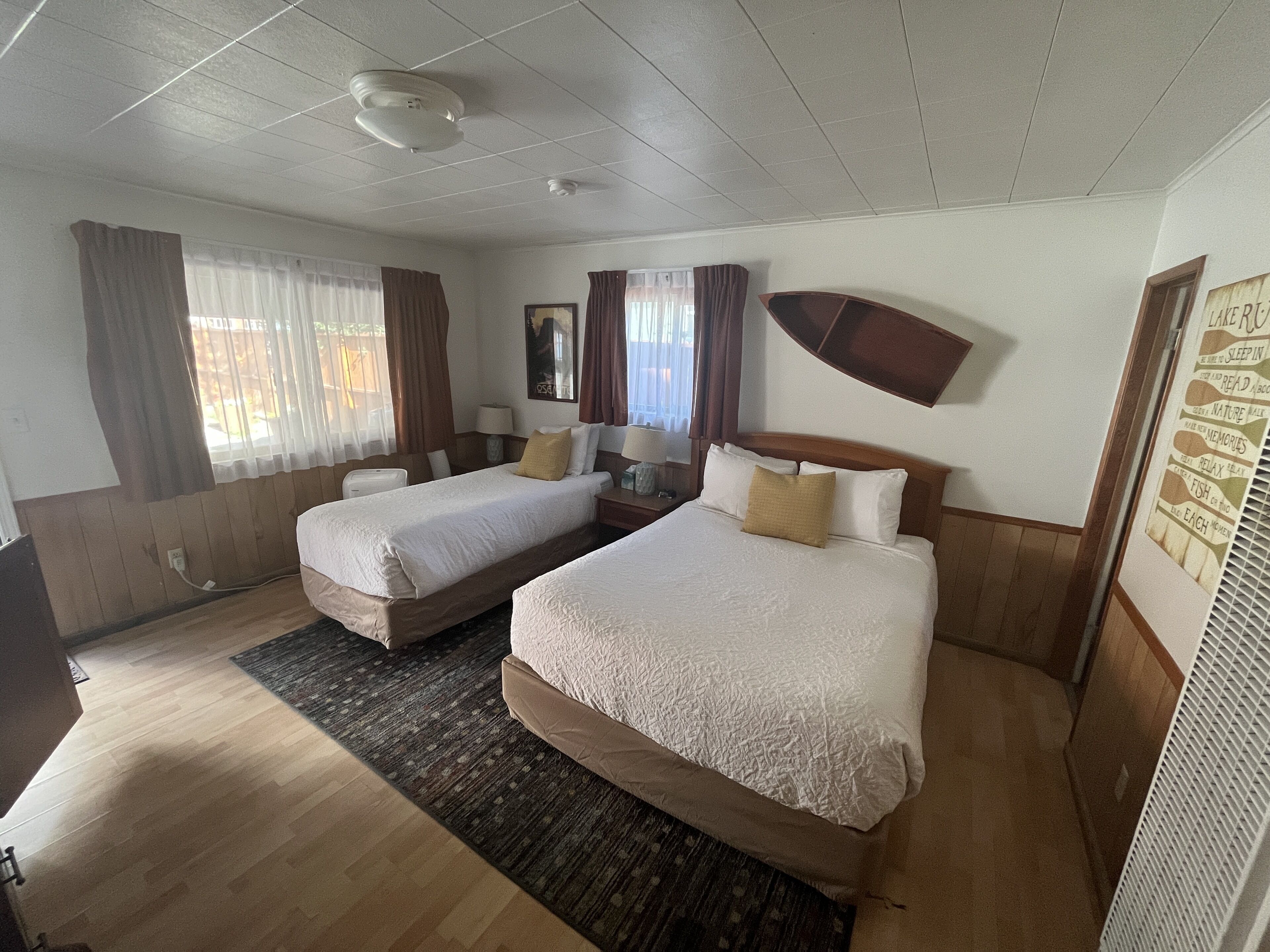 A hotel room with two beds, a wooden floor, a ceiling fan, and a wall-mounted shelf with a sign.