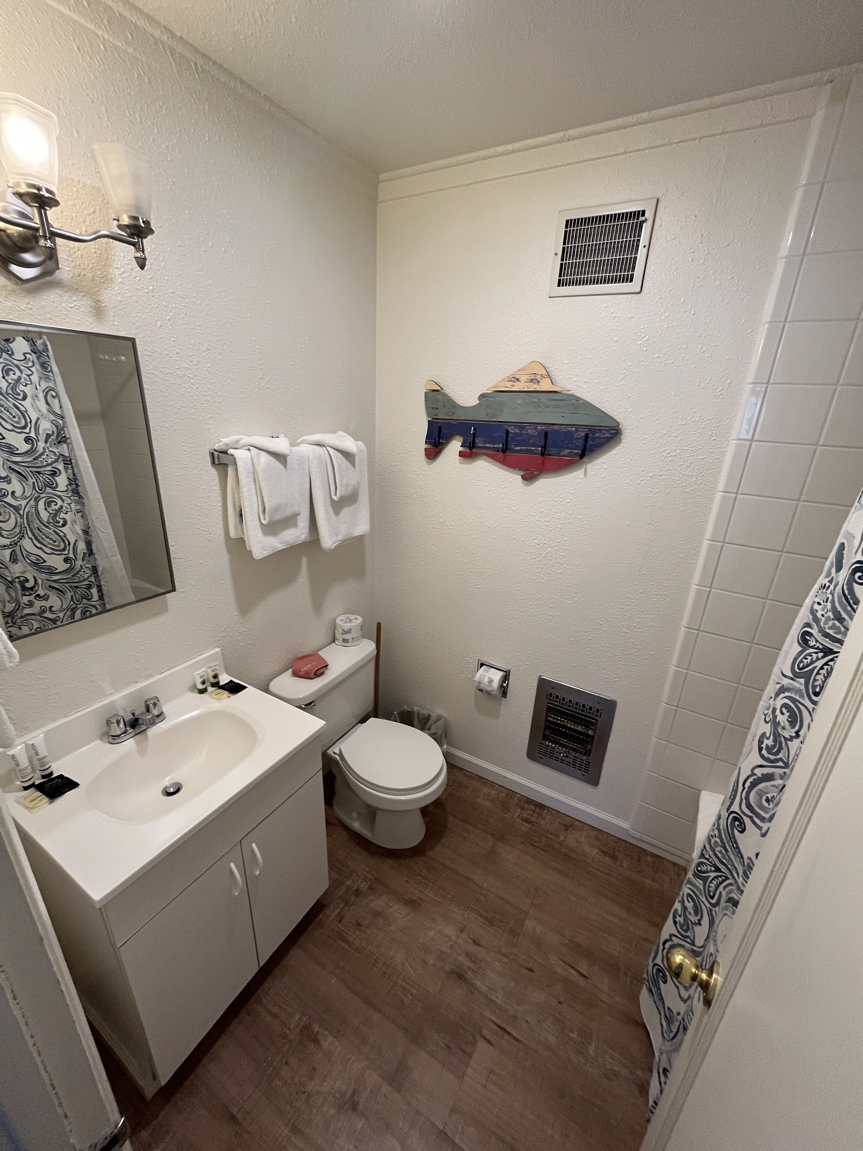Un baño con lavamanos, inodoro y ducha blancos. Hay un perchero con toallas, un espejo y un estante decorativo con forma de pez.