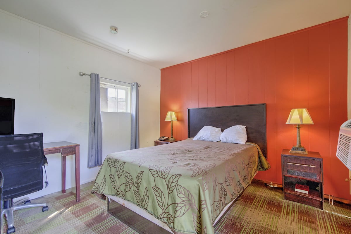 A hotel room with a bed, two bedside lamps, a desk with a chair, a window with curtains, and a wall heater.