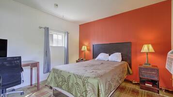 A hotel room with a bed, two bedside lamps, a desk with a chair, a window with curtains, and a wall heater.