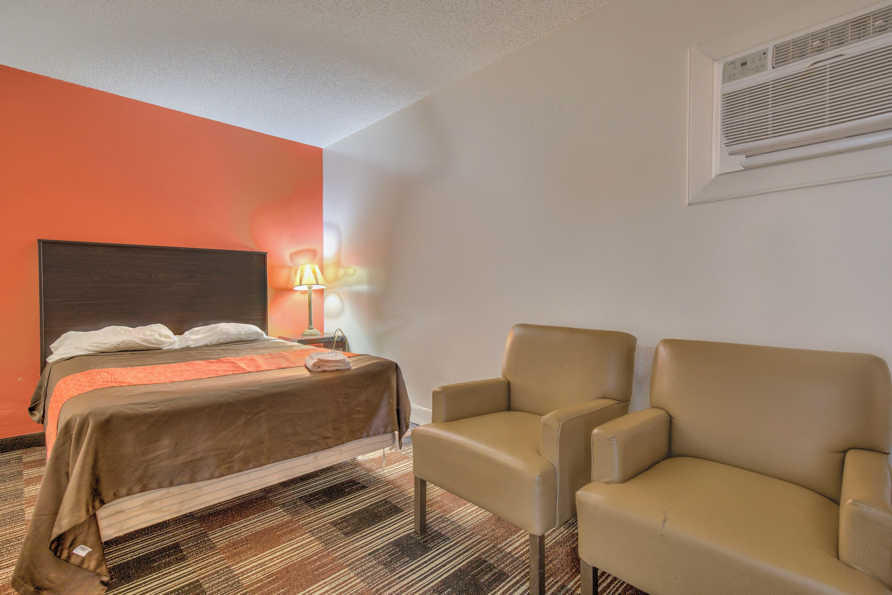 Una habitación de hotel con una cama, dos sillones, una mesita de noche, una lámpara y un equipo de aire acondicionado.