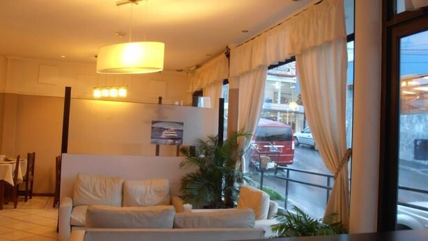 Une chambre d’hôtel avec un canapé, une table à manger et des chaises. Il y a une fenêtre avec des rideaux, et on peut voir un fourgon rouge à l’extérieur.
