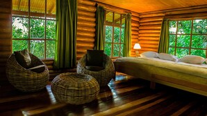 A log cabin room with a bed, two wicker chairs, a small table, and large windows with curtains. - Huasai Wasi Ecolodge (San Antonio de Cumbaza)