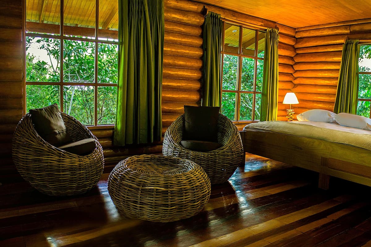 A log cabin room with a bed, two wicker chairs, a small table, and large windows with curtains.