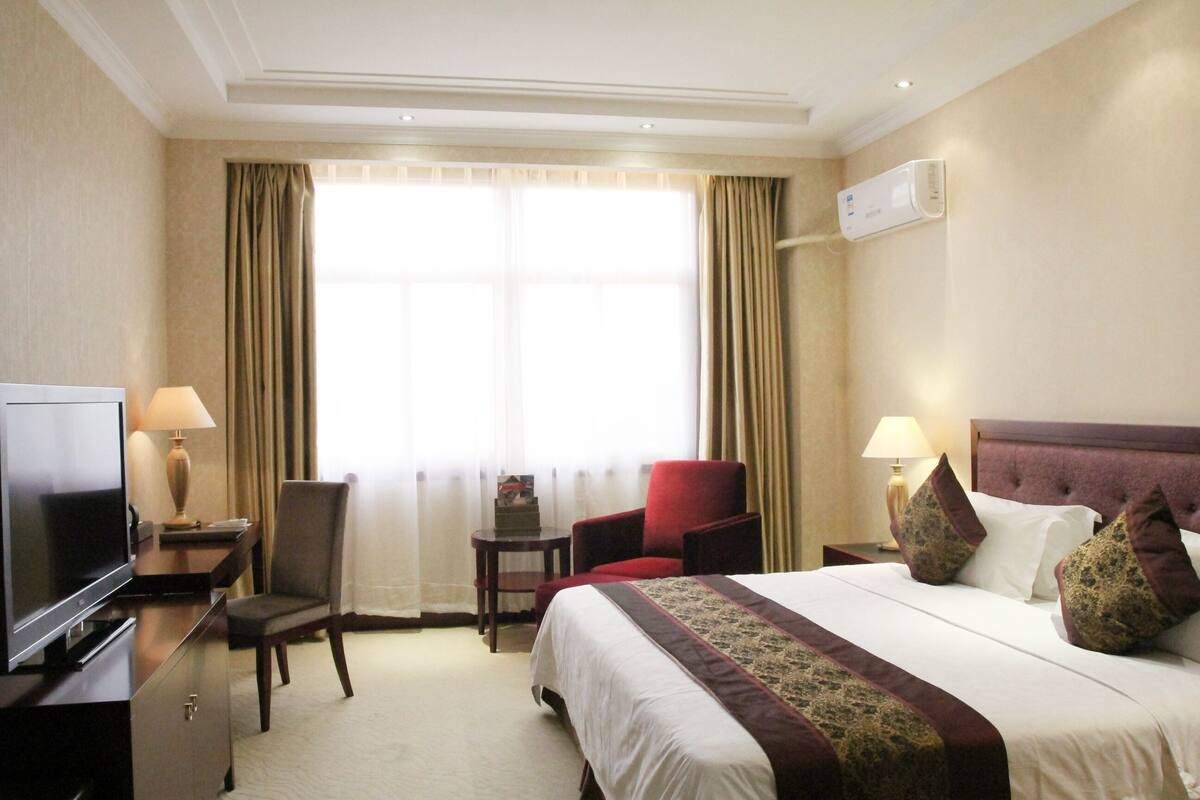 A hotel room with a bed, a TV, a desk with a lamp, a chair, and a small table.