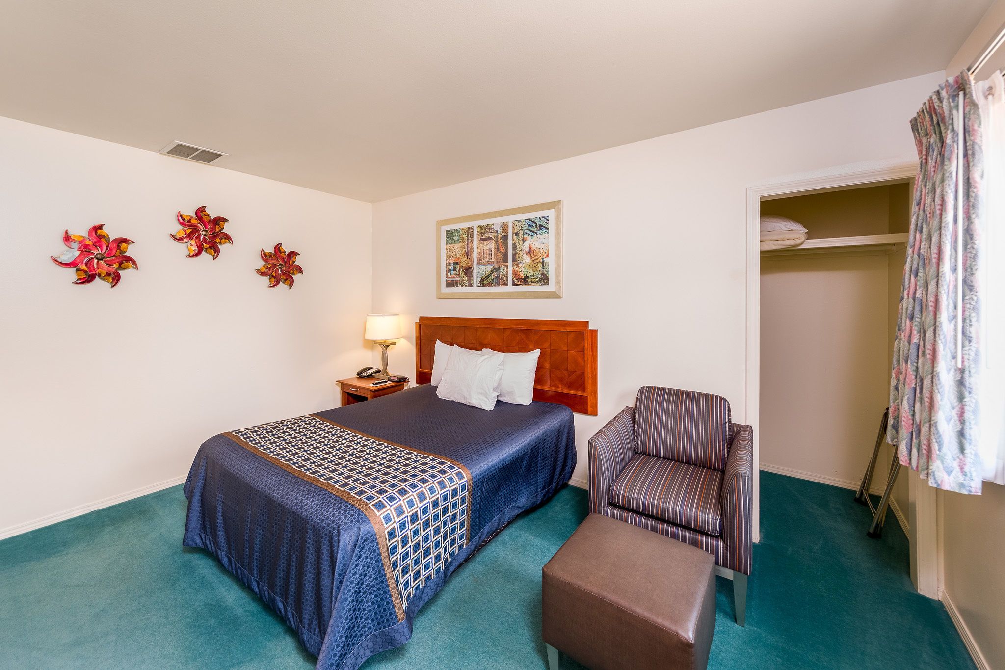 A hotel room with a large bed, a chair, a small table, a nightstand, a lamp, a painting, and a closet.
