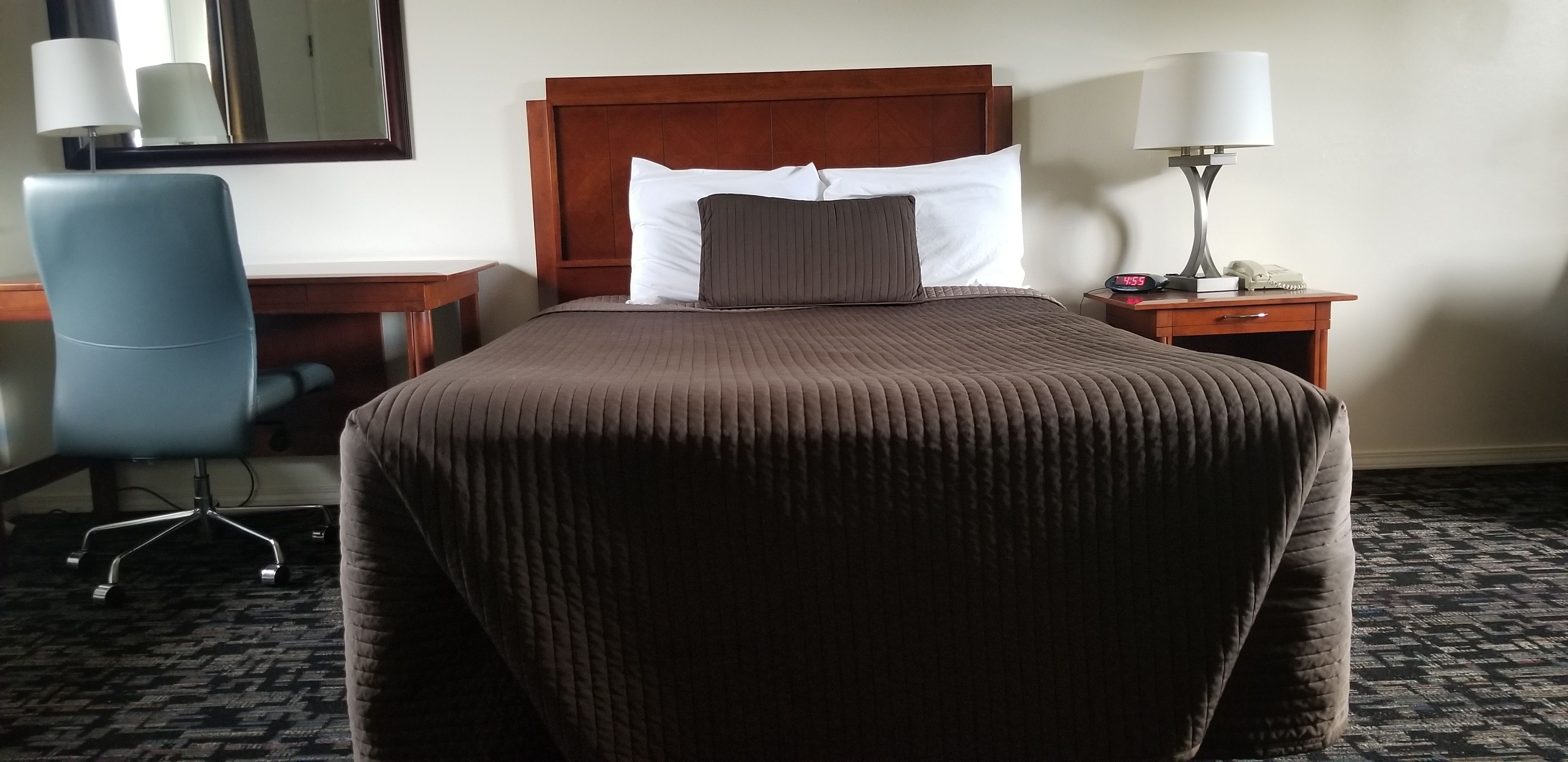 A hotel room with a large bed, a wooden headboard, a desk with a chair, a nightstand with a lamp, and a mirror on the wall.