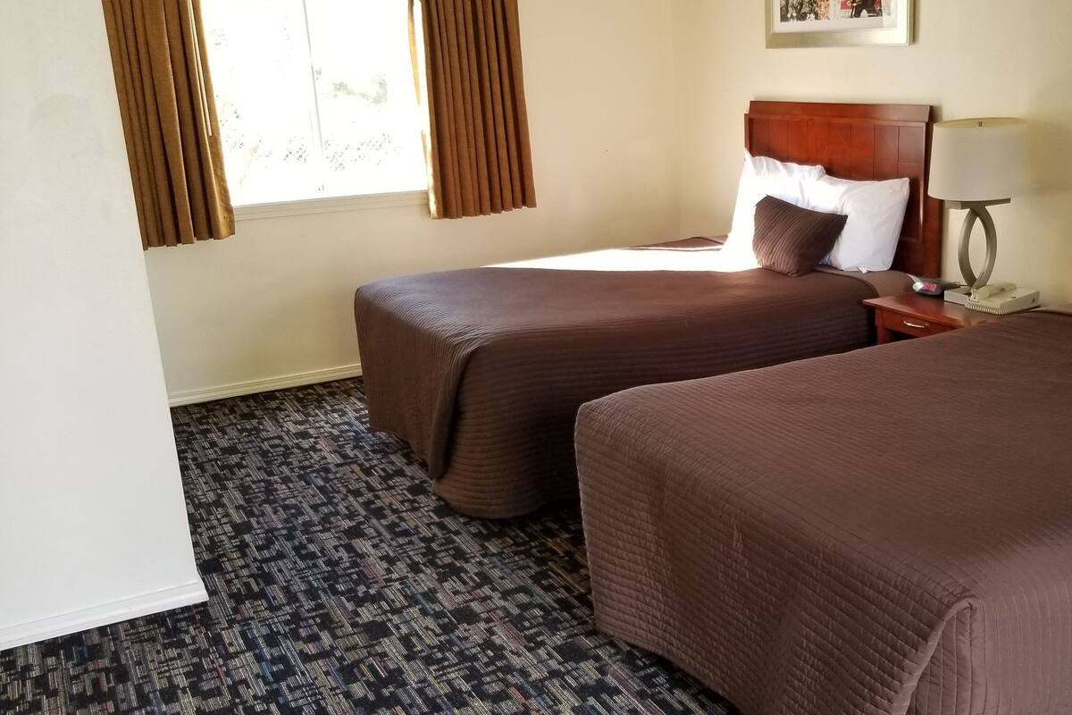 A hotel room with two beds, a nightstand, a lamp, and a window with curtains.
