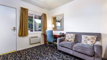 A hotel room with a gray sofa, a blue office chair, a desk with a lamp, a window with curtains, and a radiator.