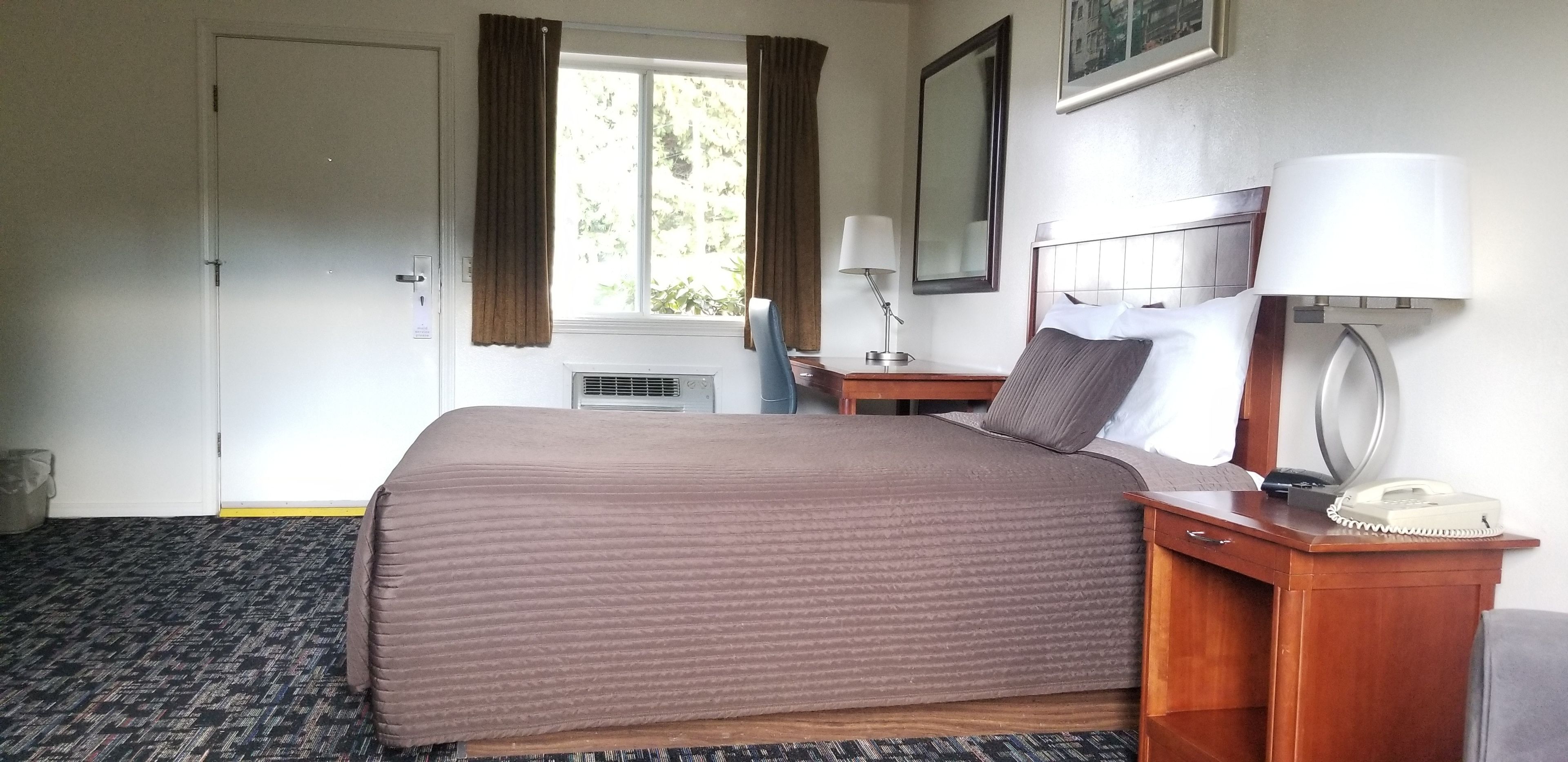 A hotel room with a bed, bedside table, lamp, phone, and a window with curtains.