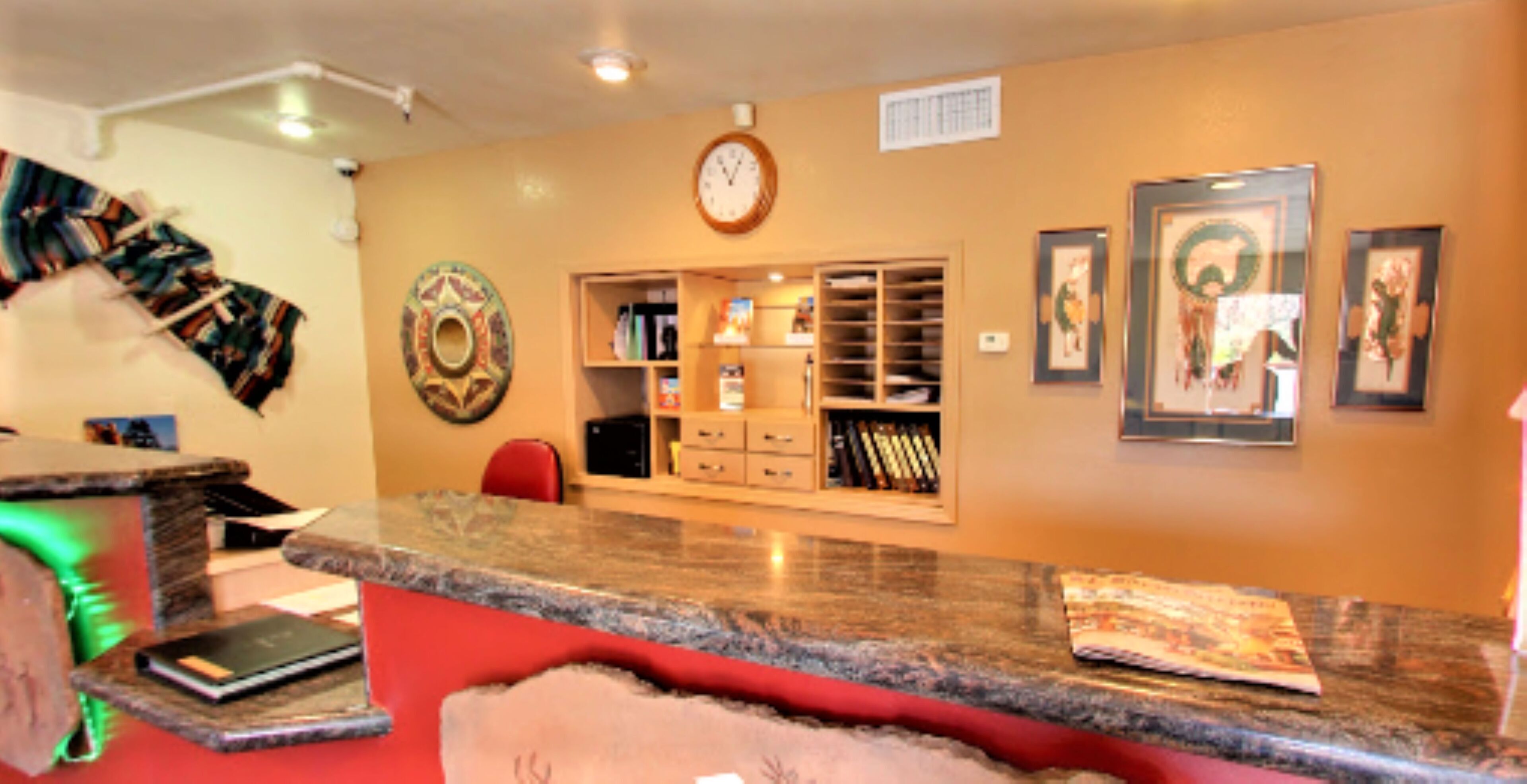 A room with a kitchen counter, a clock, shelves with books, and framed pictures on the wall.
