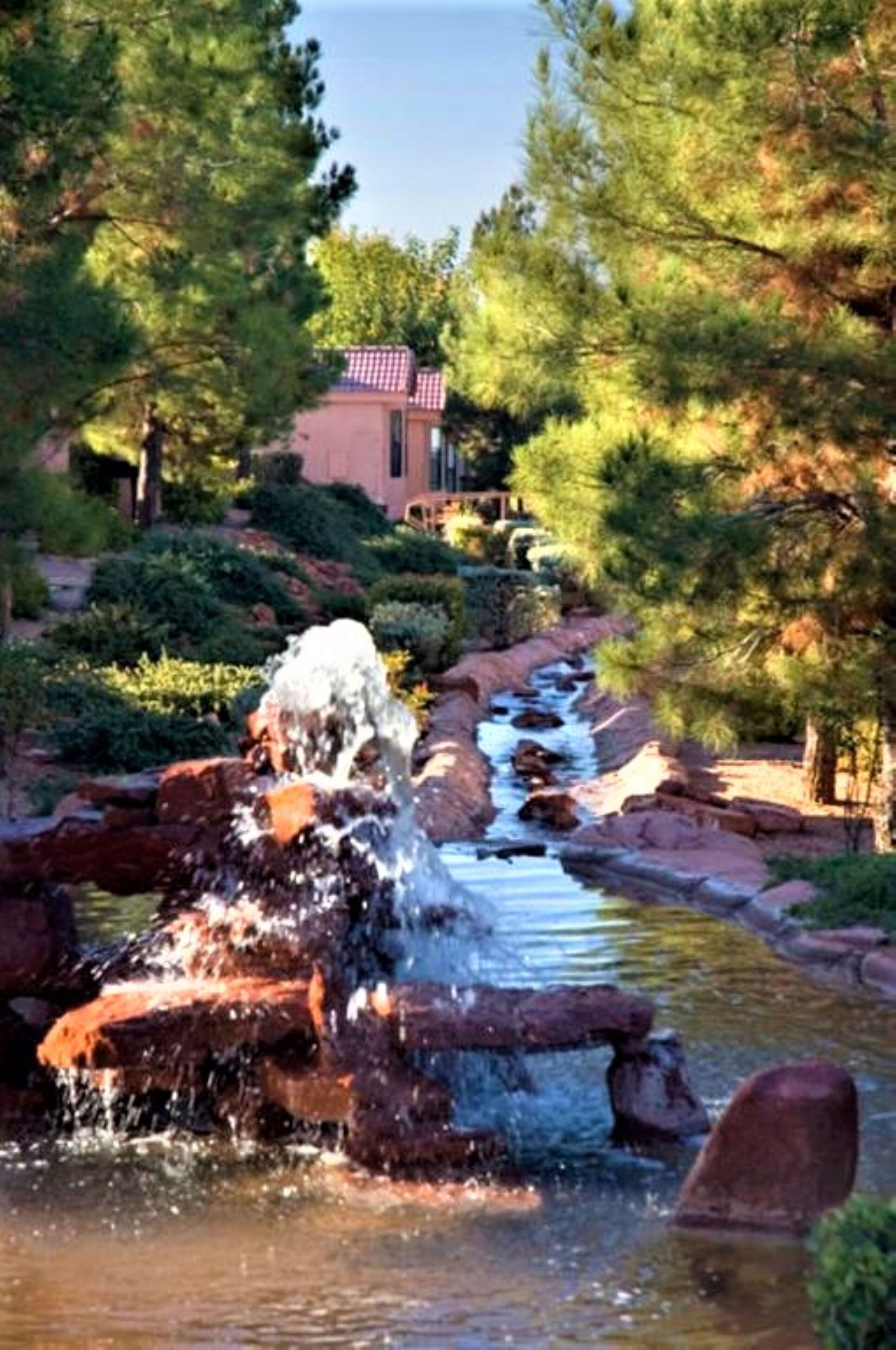 A serene outdoor setting with a small waterfall, surrounded by lush greenery and a building with a red roof.