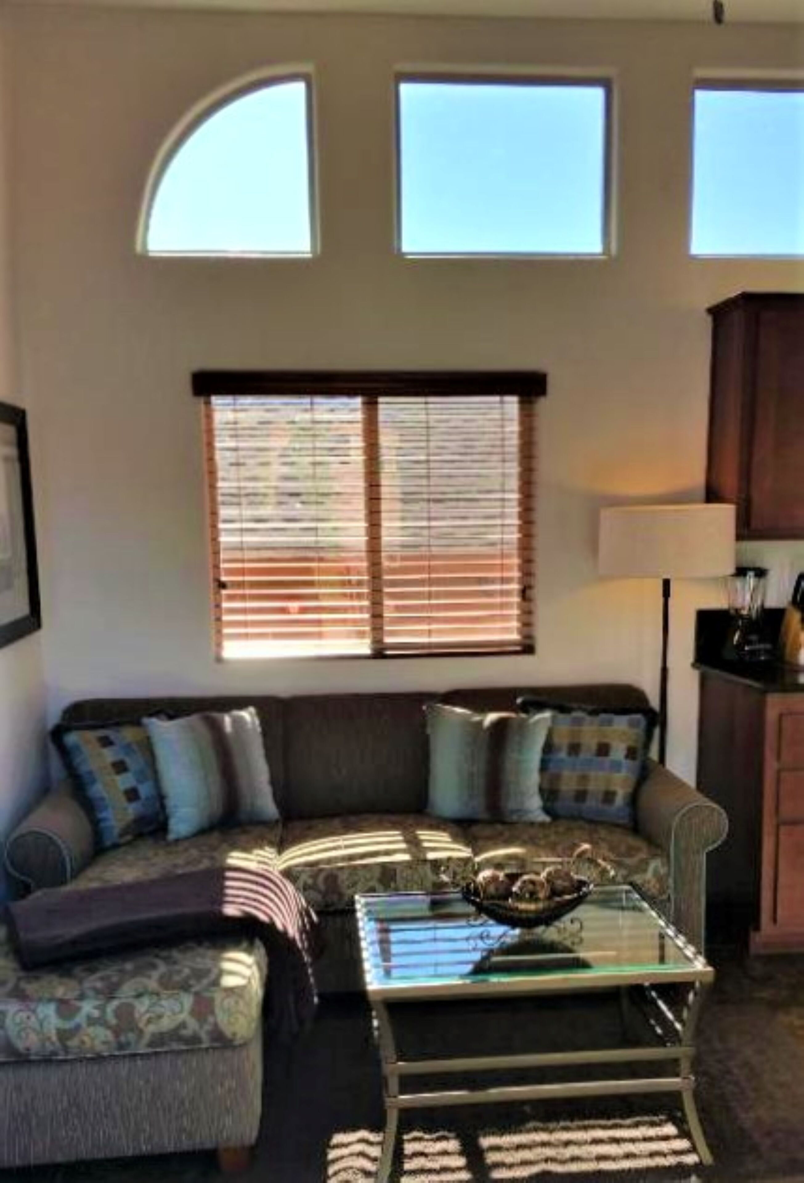 A living room with a sofa, a glass coffee table, and a lamp. There are two windows with blinds and a curved ceiling.