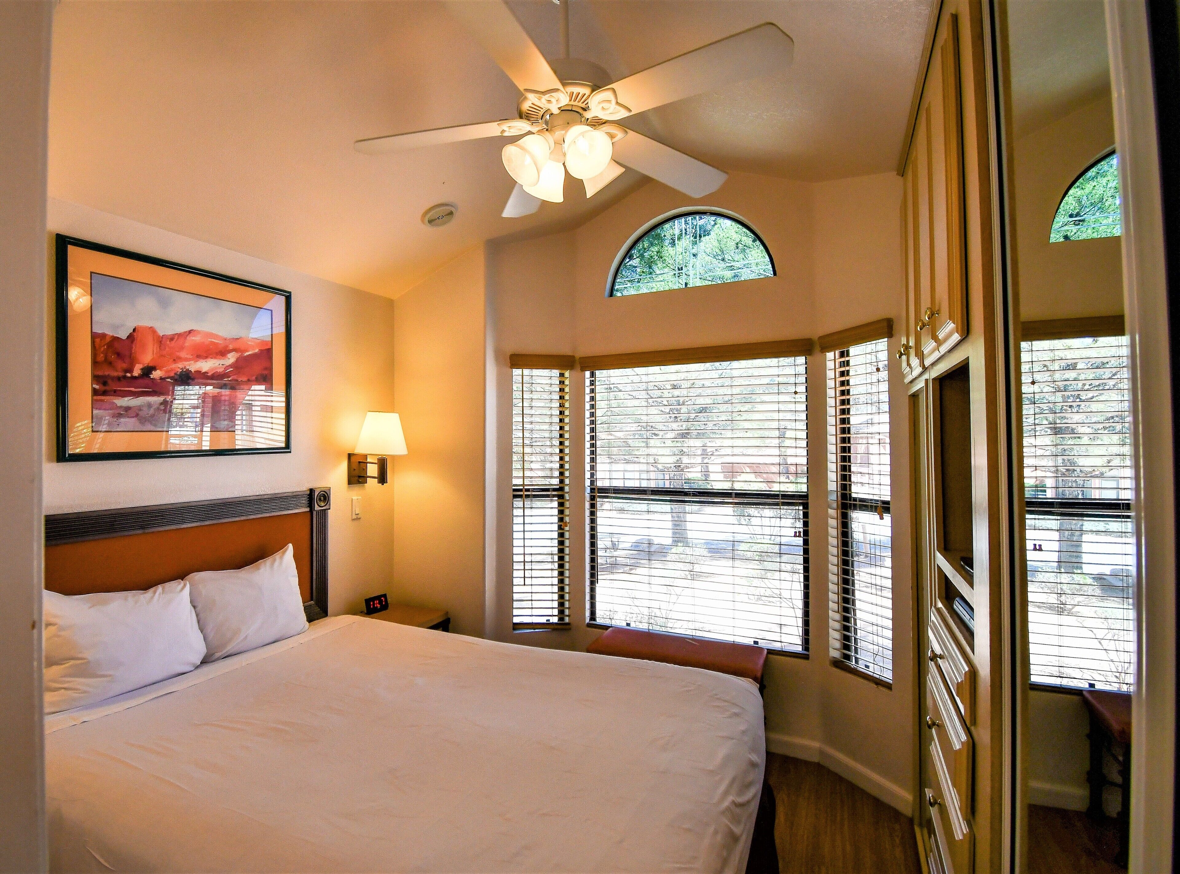 Une chambre d’hôtel avec un lit, un ventilateur de plafond, un tableau, une lampe et de grandes fenêtres équipées de stores.
