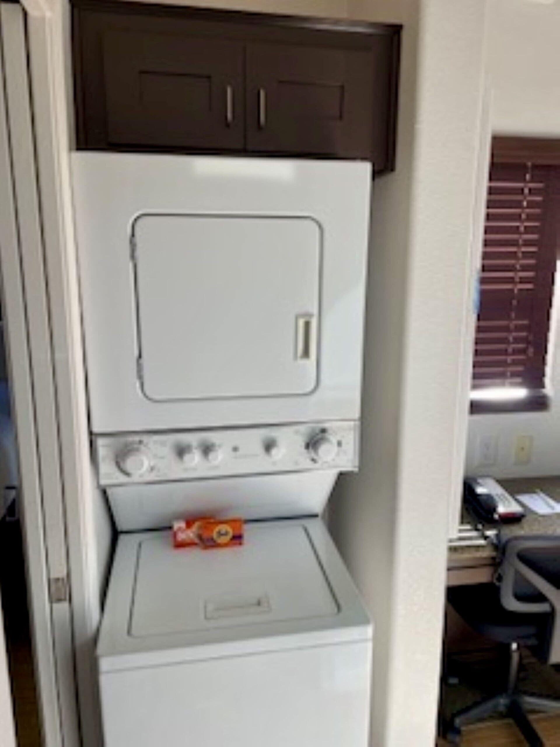Una cocina compacta con estufa blanca, horno pequeño y microondas. Hay una ventana con persianas, una silla y un escritorio con una computadora.