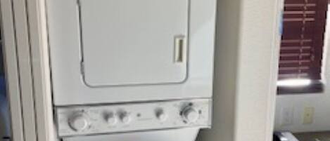A compact kitchen with a white stove, a small oven, and a microwave. There is a window with blinds, a chair, and a desk with a computer.