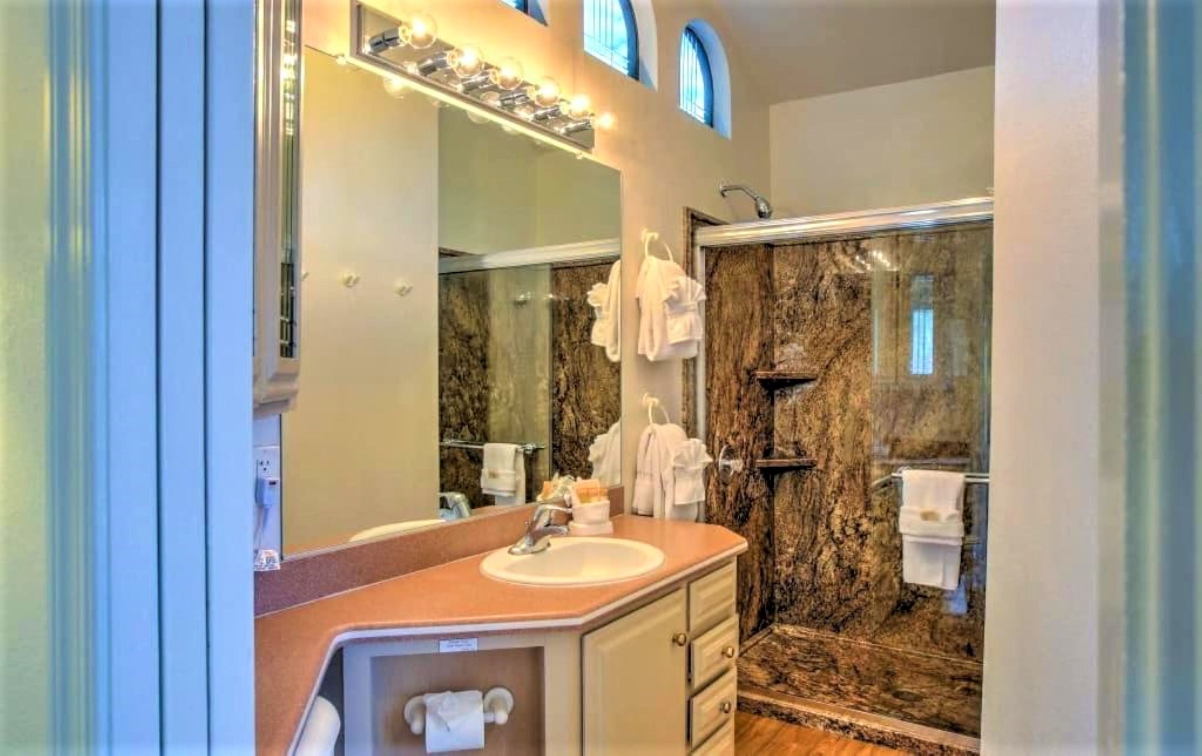 A bathroom with a marble shower, a sink, and a large mirror with lights.