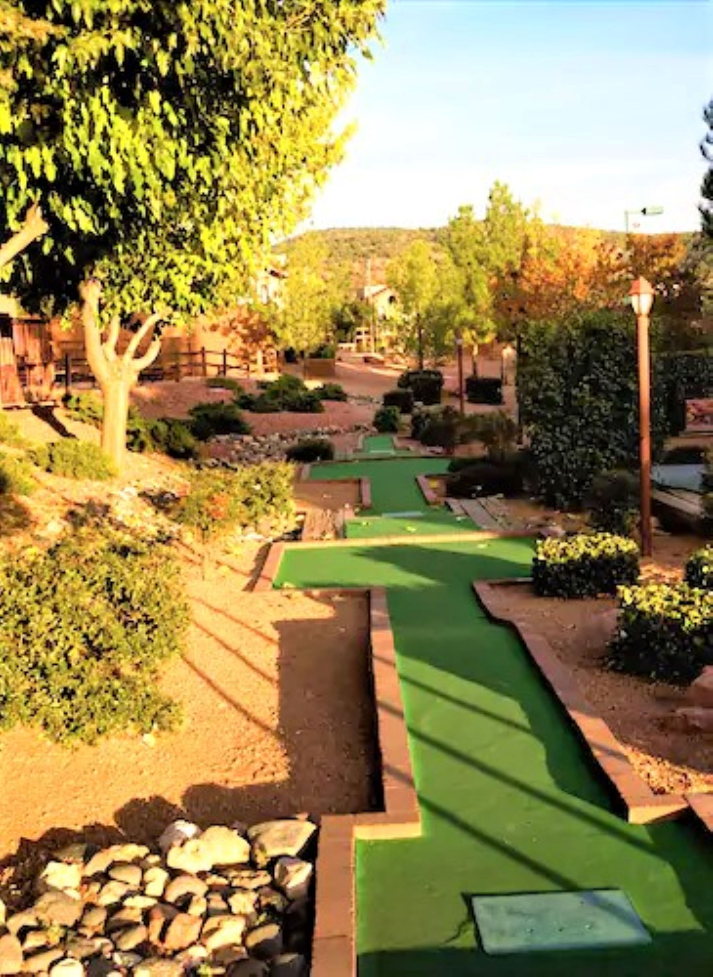 A miniature golf course with a green putting surface, surrounded by landscaped areas and trees.