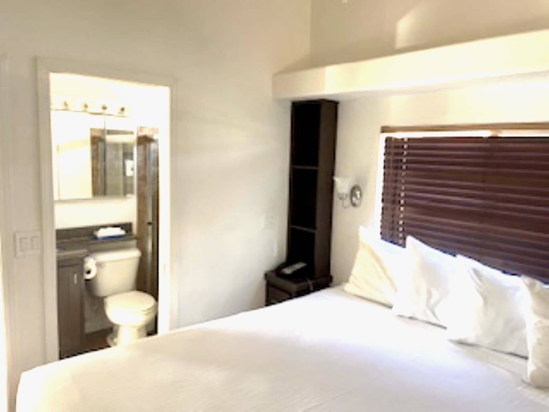Habitación de hotel con cama, mesita de noche, espejo, ventana con persianas y baño con inodoro y lavamanos.