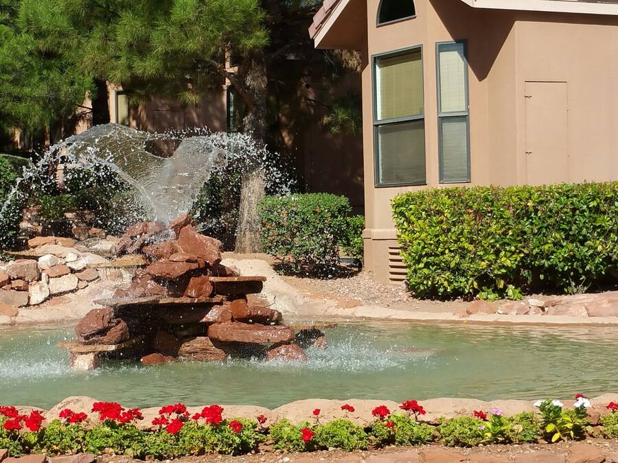 A fountain surrounded by a stone border and red flowers, with a building and hedges in the background.